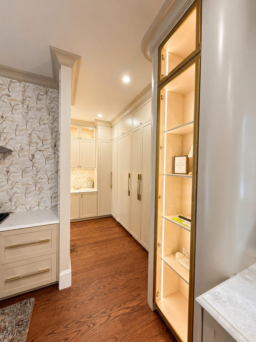 Hallway with a tall gold-framed illuminated glass display cabinet beside floral wallpaper and white cabinetry