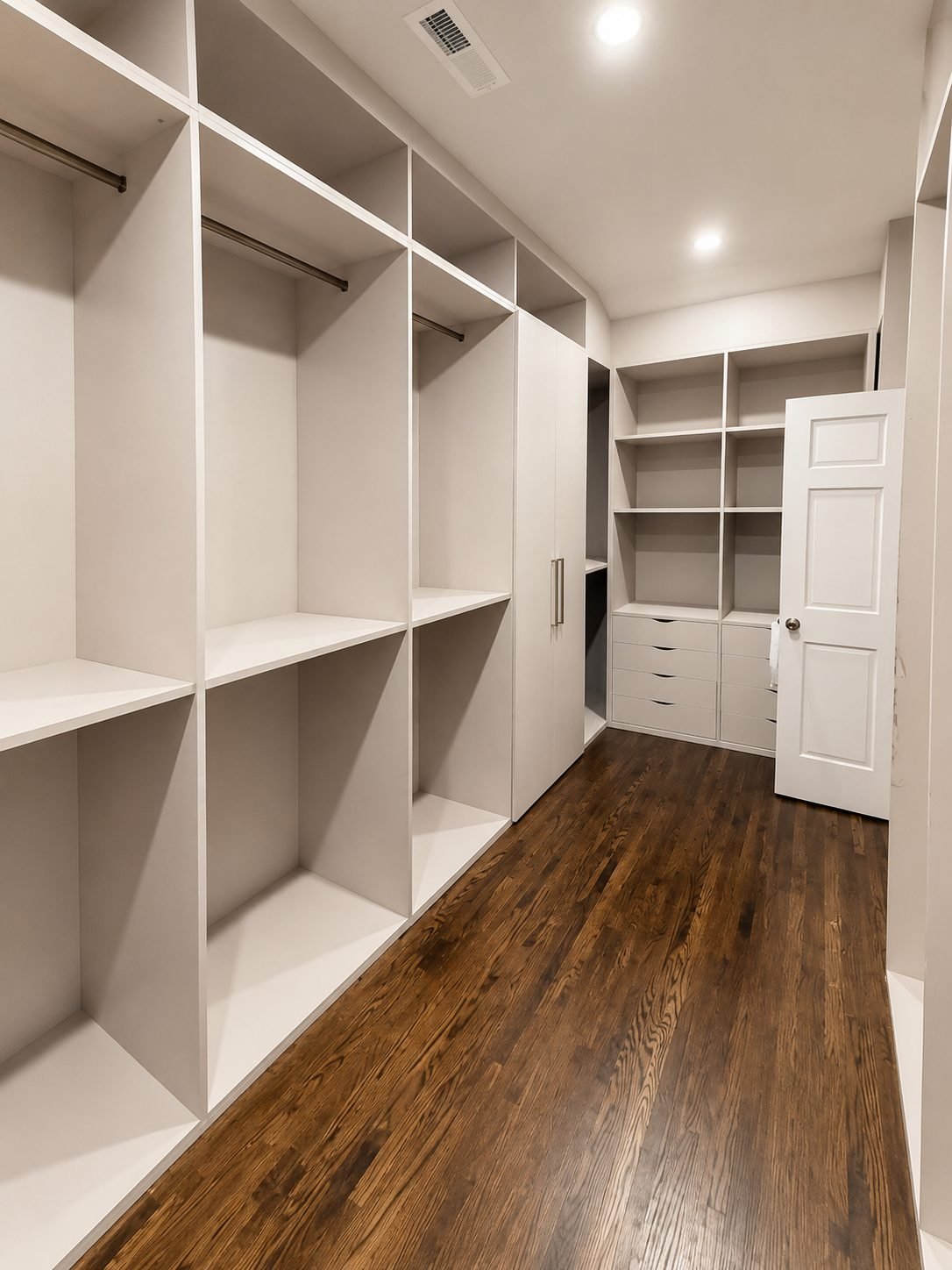 Empty greige walk-in closet with open hanging bays, drawers and a door to the hallway, dark wood floor