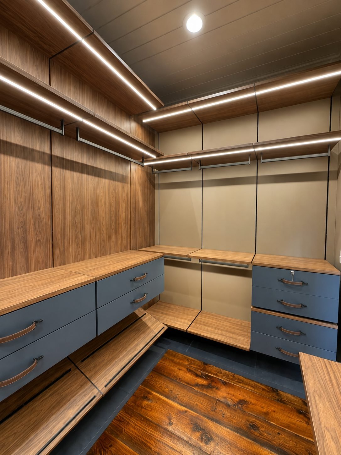 Walnut walk-in closet with slate-blue drawers, leather pulls, linear LED shelf lighting and a reclaimed reddish-wood floor inset